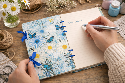 Person writing in a journal with a floral cover on a wooden table.